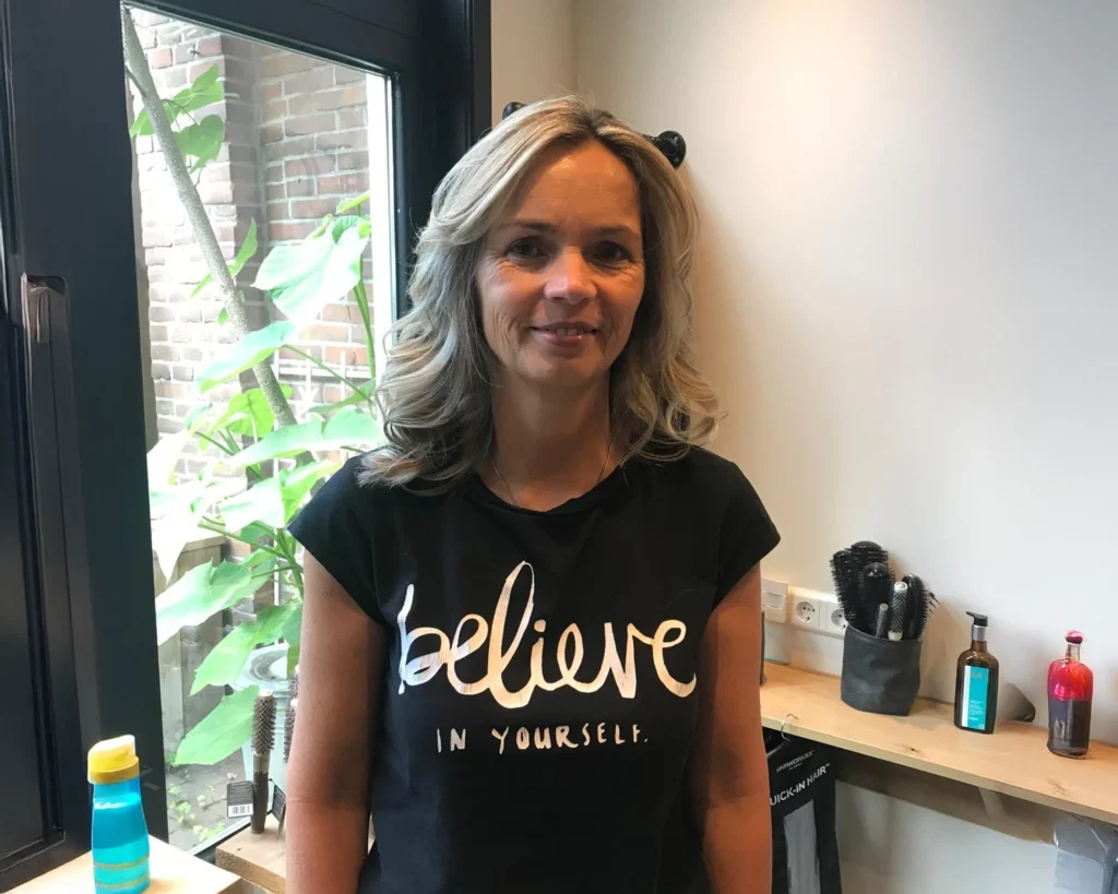 Een vrouw staat binnen met krullend blond haar, draagt een zwart t-shirt met de tekst believe in yourself. Achter haar staat een plank met flessen en een raam met groene planten buiten. - massagesalon Mari-Sage Nieuwegein